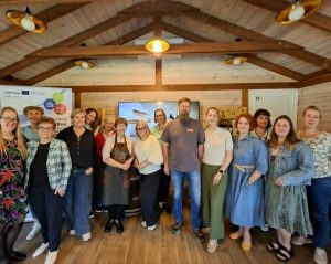 Group photo of participants at a workshop in a wooden cabin setting, featuring diverse individuals engaged in community initiatives. The background includes banners and a screen displaying relevant information, highlighting collaboration in local projects.