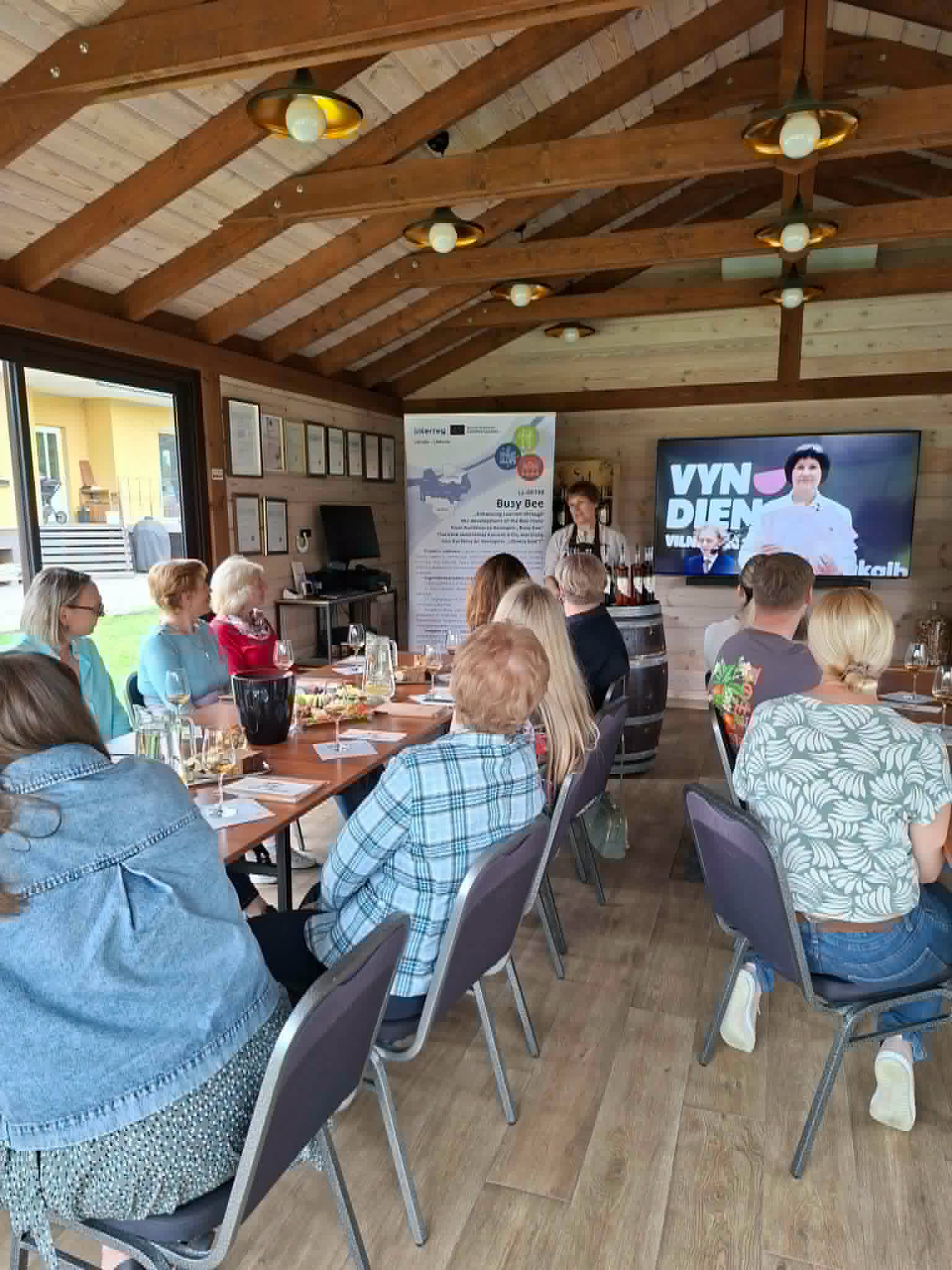 A group of attendees sitting at a wooden table in a cozy venue, listening to a presentation on winemaking. A screen displays the text "VYN DIEN," while a banner promoting a local business event is visible in the background. The setting features wooden beams and a warm atmosphere, with glasses and plates of food on the table.