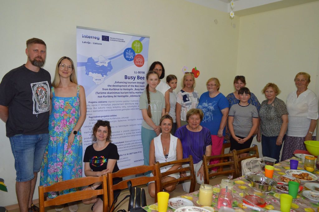Group photo of participants in the Busy Bee project, promoting tourism development from Kuršėnai to Ventspils. The image features a diverse group of individuals posing in front of an informational banner, surrounded by a table with colorful drinks and food. This initiative aims to enhance community engagement and local tourism.