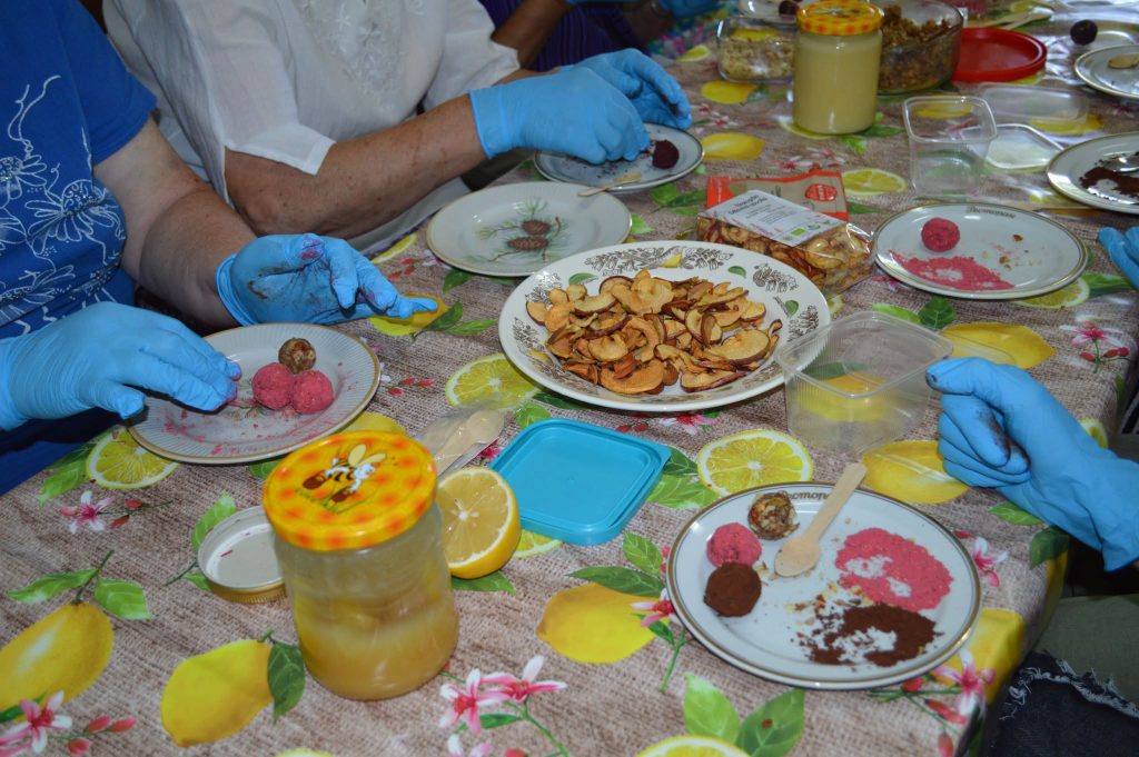 Participants in a food preparation activity wearing blue gloves create colorful snacks on a table covered with a lemon-patterned cloth. The scene includes plates of pink and chocolate-coated treats, dried fruit chips, and jars of ingredients, showcasing a vibrant and engaging culinary experience.