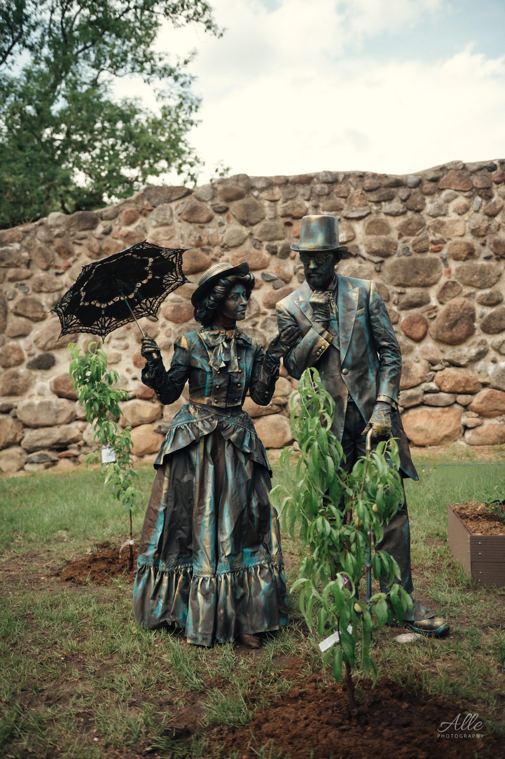 A pair of street performers dressed in vintage attire, including a woman with a parasol and a man in a top hat, stand among young trees in a garden setting, with a stone wall in the background. A pair of street performers dressed in vintage attire, including a woman with a parasol and a man in a top hat, stand among young trees in a garden setting, with a stone wall in the background.