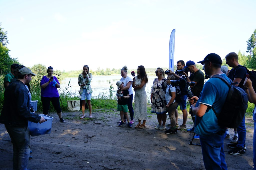 Group of people gathered at the Ruģeļi Wetland in Daugavpils, Latvia, watching the release of turtles into the wild. The scene includes both adults and children, with some participants taking photos and videos, highlighting community engagement in wildlife conservation efforts.