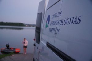 A researcher from the Latvian Institute of Hydrology stands by a calm lake at dusk, next to a small boat and a van displaying the institute's logo.