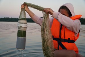 A person wearing a life jacket collects water samples using a filtration device in a lake at sunset, demonstrating environmental research practices.