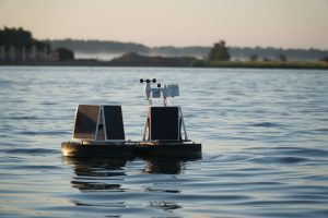 Autonomous water monitoring buoy equipped with solar panels, floating on calm water during sunrise, with a background of misty shoreline vegetation.