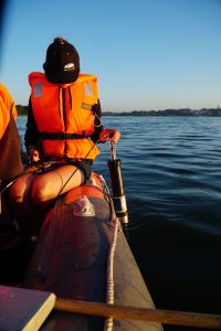 Person in an orange life jacket sitting in a boat, using a device with a cable, on a calm water surface during sunset.