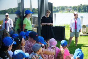 Group of children wearing blue caps listening attentively to two women speaking outdoors by a lake. The scene captures an educational moment in a sunny environment, highlighting community engagement and youth activities.