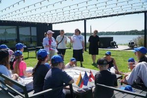Group of children wearing blue caps seated outdoors, listening to three women presenting at a lakeside venue. Flags representing Lithuania, the European Union, and Latvia are displayed on a table. The scene captures a summer educational event with a focus on community engagement and cultural exchange.