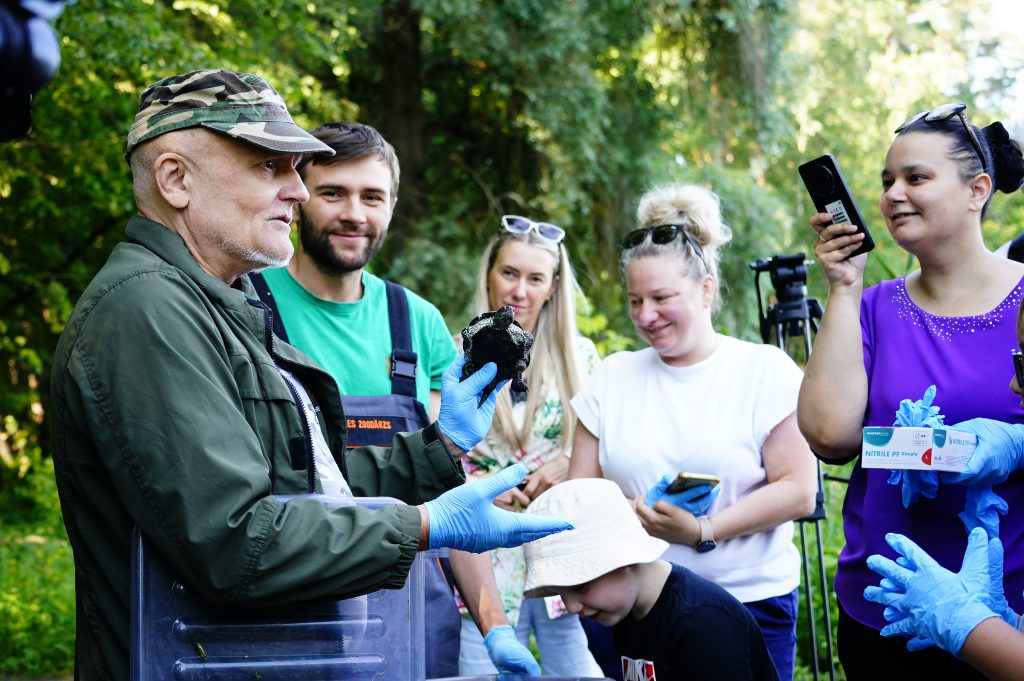 Group of people, including a man in a camouflage cap, gathering at Ruģeļi Wetland in Daugavpils, Latvia, to participate in the release of turtles into the wild. The man is holding a turtle while others observe and take photos, highlighting community involvement in wildlife conservation efforts.