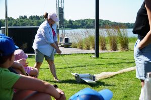 A woman demonstrates the use of a fishing net on a sunny day by the lake, while children observe attentively. The scene highlights outdoor educational activities related to fishing and marine life.