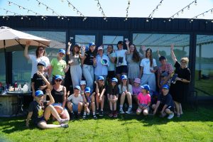 Group photo of children and adults at a summer camp, smiling and waving, with blue caps and a sunny outdoor setting featuring string lights and a building in the background.