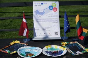 Image of a promotional display for the "Lakes Go Digital" project, featuring flags of Latvia, the European Union, and Lithuania. The display includes information about the project's goals, budget, and timeline, alongside handmade decorative plates illustrating themes related to lakes and nature. The setting is outdoors on a sunny day, with green grass in the background.