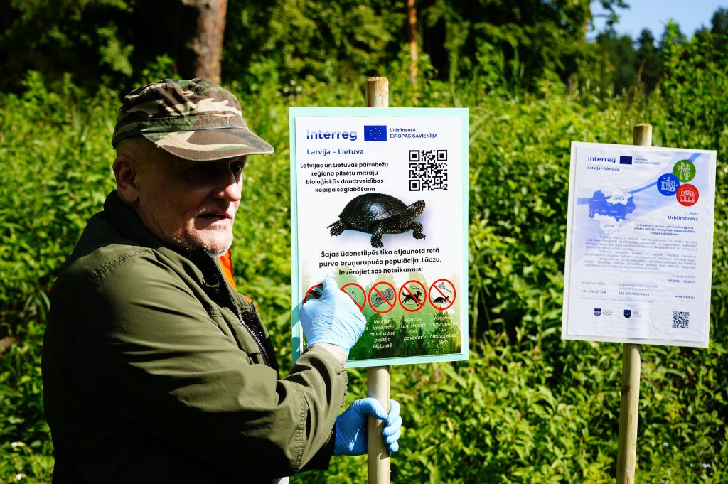 An individual in a camouflage cap holds a sign about turtle conservation at the Ruģeļi Wetland in Daugavpils, Latvia, surrounded by lush greenery. The sign provides guidelines for protecting a rare turtle population and features a QR code for additional information.