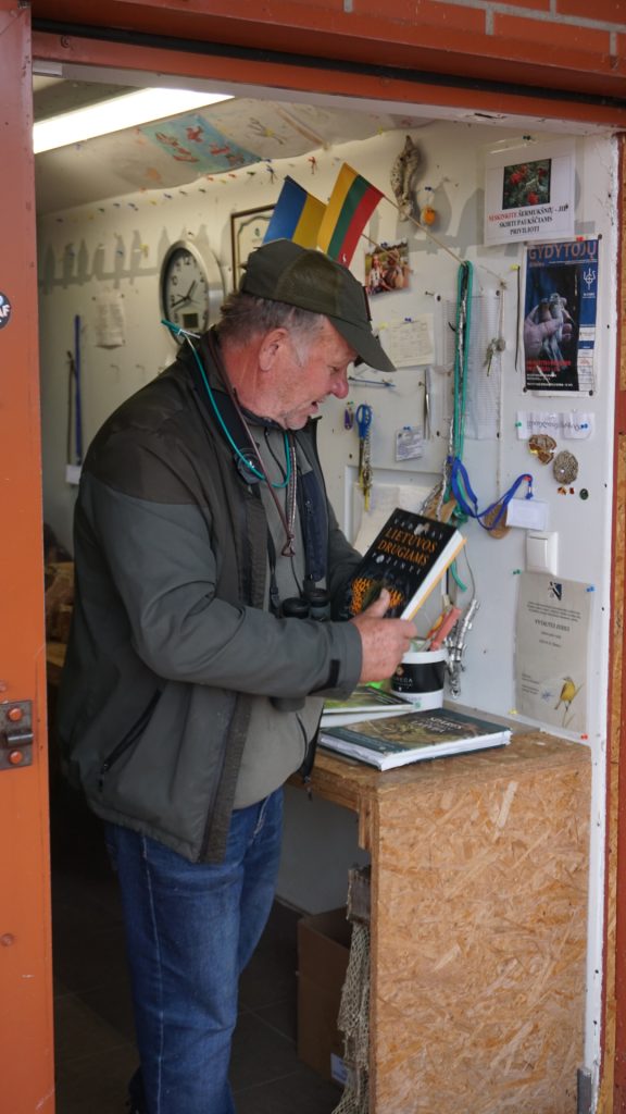 A man holding a book titled "Lietuvos Drugiais" stands in a small shop adorned with Lithuanian flags and various nature-related materials, as part of an initiative to explore BIRDIE results in the Klaipėda region.