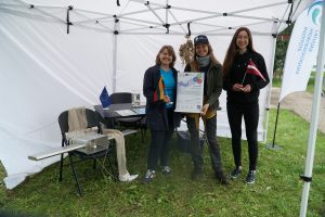 Three women stand under a white tent, promoting environmental awareness. They hold flags from Germany and Latvia, alongside a poster detailing their project. A table displays educational materials and a water sample. The setting is grassy, indicating an outdoor event focused on ecology and sustainability.