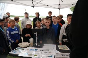 A group of children attentively listening to a presentation at an outdoor educational event, with informational materials displayed on a table under a tent. The background features a lake and greenery, highlighting a focus on environmental education.