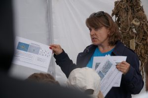 A woman in a blue shirt and jacket passionately explains marine life concepts to an audience, holding informational sheets with images and descriptions. The setting is a tent, with a background of marine materials, highlighting an educational event focused on ocean conservation and marine biology.
