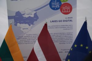 Flags of Lithuania, Latvia, and the European Union in front of a promotional poster for the "Lakes Go Digital" project, which focuses on water quality monitoring in the Baltic region. The poster features a map highlighting Latvia and Lithuania, emphasizing digital solutions for environmental management.
