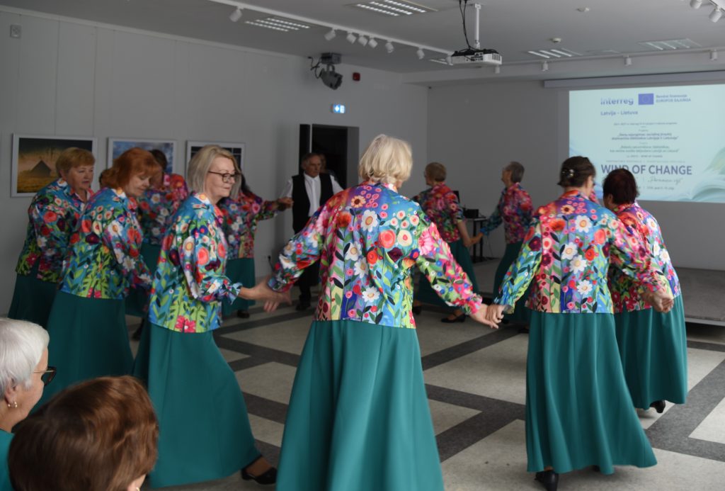 A group of women in colorful floral jackets and long green skirts dance together in a vibrant indoor setting, celebrating community and cultural expression at an event titled "Wind of Change."