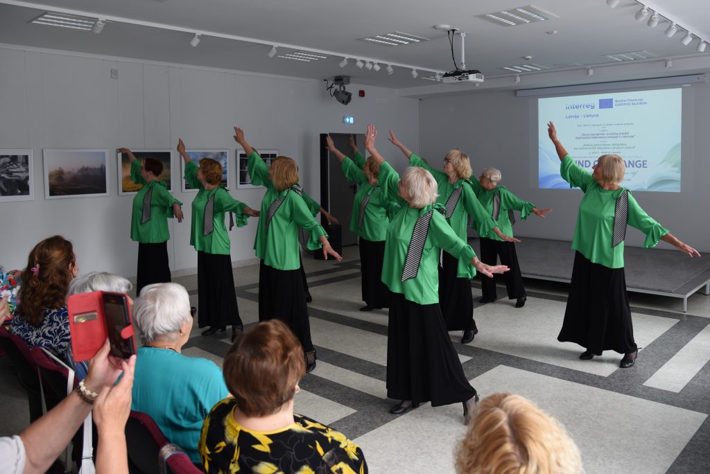 A group of elderly women in green outfits perform a dance on stage during a cultural event, with an audience seated in the foreground. The background features photographs and a presentation screen, highlighting the community engagement aspect of the event.