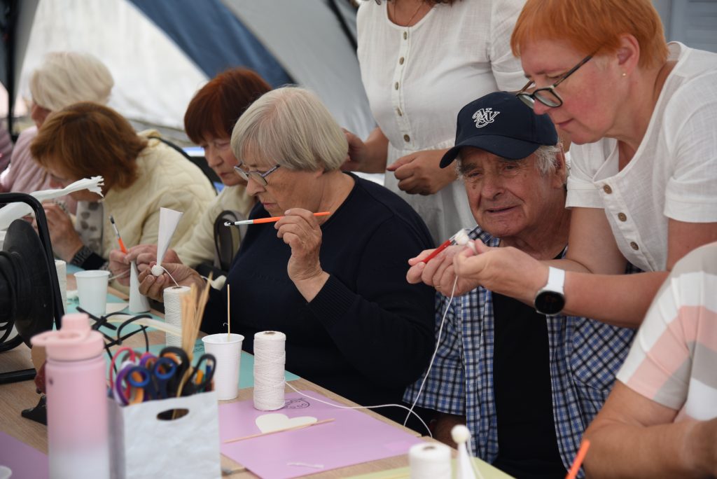 Elderly participants engaged in a crafting workshop, focused on creating handmade projects using various materials. The scene highlights collaboration and creativity, with individuals using tools like scissors, paint, and thread, set in a tented environment.