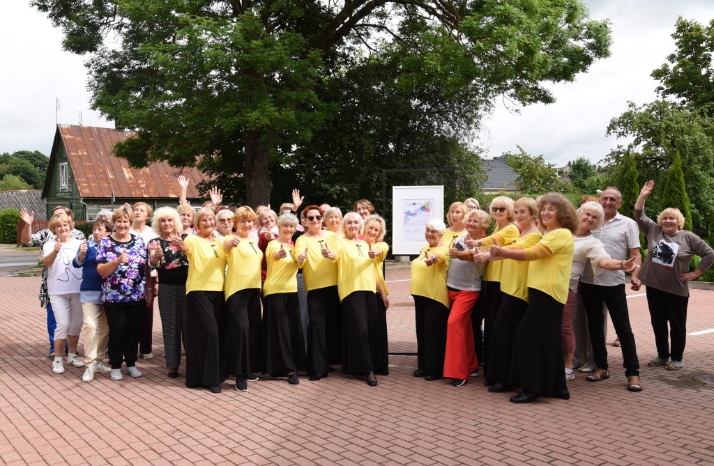 Group of elderly women in matching yellow tops, smiling and posing outdoors, with some giving thumbs up and waving. The background features a tree and a building with a rusty roof, creating a vibrant community atmosphere.