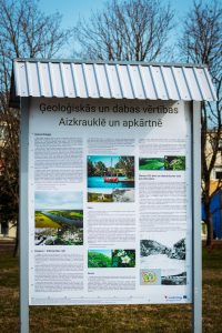 Informative sign detailing the geological and natural values of Aizkraukle, featuring sections on geomorphology, flora, and fauna, with accompanying images. The background includes trees and a clear sky. Informative sign detailing the geological and natural values of Aizkraukle, featuring sections on geomorphology, flora, and fauna, with accompanying images. The background includes trees and a clear sky.