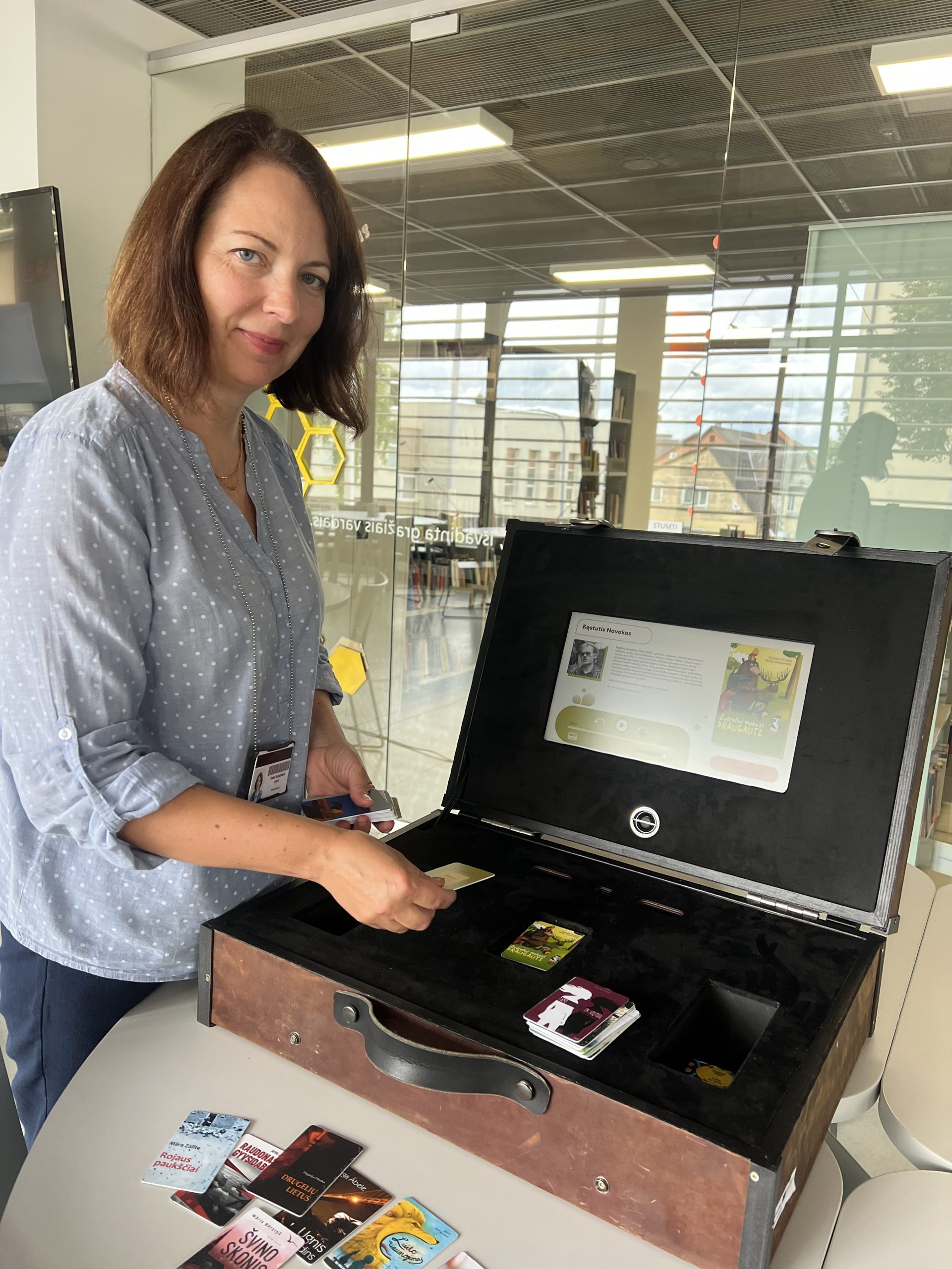 A woman interacts with an interactive book suitcase, exploring literature through cards displayed on a table. The suitcase features a digital screen and various book covers, set in a modern library environment. A woman interacts with an interactive book suitcase, exploring literature through cards displayed on a table. The suitcase features a digital screen and various book covers, set in a modern library environment.