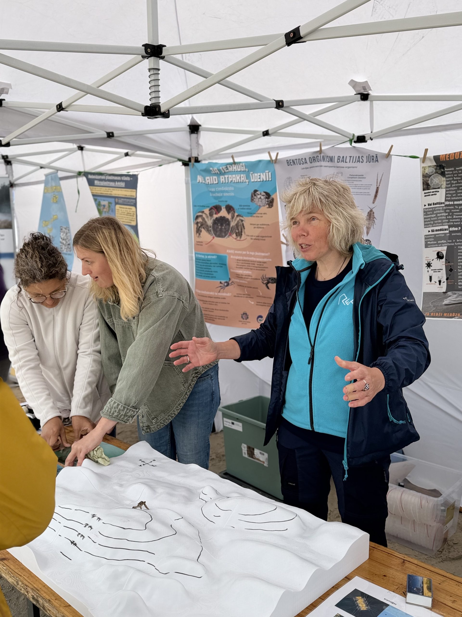 Three women engage in a hands-on educational activity at a booth, discussing marine life and conservation. The table displays a tactile model of aquatic organisms, while informative posters about benthic organisms and local marine ecology are visible in the background. Three women engage in a hands-on educational activity at a booth, discussing marine life and conservation. The table displays a tactile model of aquatic organisms, while informative posters about benthic organisms and local marine ecology are visible in the background.