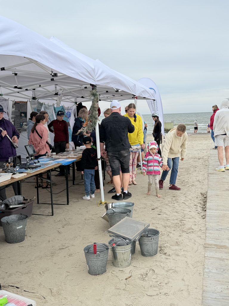 A group of people engaging in activities at a beach event, with a tent displaying information and materials. Families and children are participating, surrounded by buckets and tools on the sandy shore. The ocean is visible in the background under a cloudy sky. A group of people engaging in activities at a beach event, with a tent displaying information and materials. Families and children are participating, surrounded by buckets and tools on the sandy shore. The ocean is visible in the background under a cloudy sky.