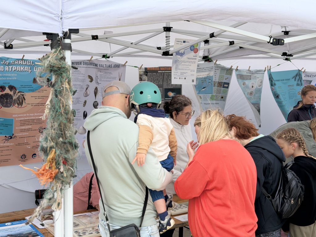 Visitors engaging with educational displays at a marine conservation booth, featuring posters about Baltic Sea organisms and environmental awareness. A parent holds a child while others examine materials and interact with staff. Visitors engaging with educational displays at a marine conservation booth, featuring posters about Baltic Sea organisms and environmental awareness. A parent holds a child while others examine materials and interact with staff.