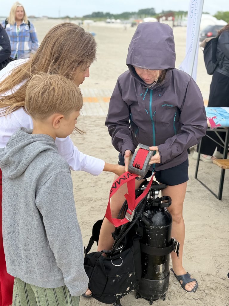 A group of three people, including a child, engaged in a hands-on diving equipment demonstration on the beach. One woman is explaining the scuba gear while another is holding a tank and accessories. The sandy beach and a cloudy sky set the scene for this educational outdoor activity. A group of three people, including a child, engaged in a hands-on diving equipment demonstration on the beach. One woman is explaining the scuba gear while another is holding a tank and accessories. The sandy beach and a cloudy sky set the scene for this educational outdoor activity.