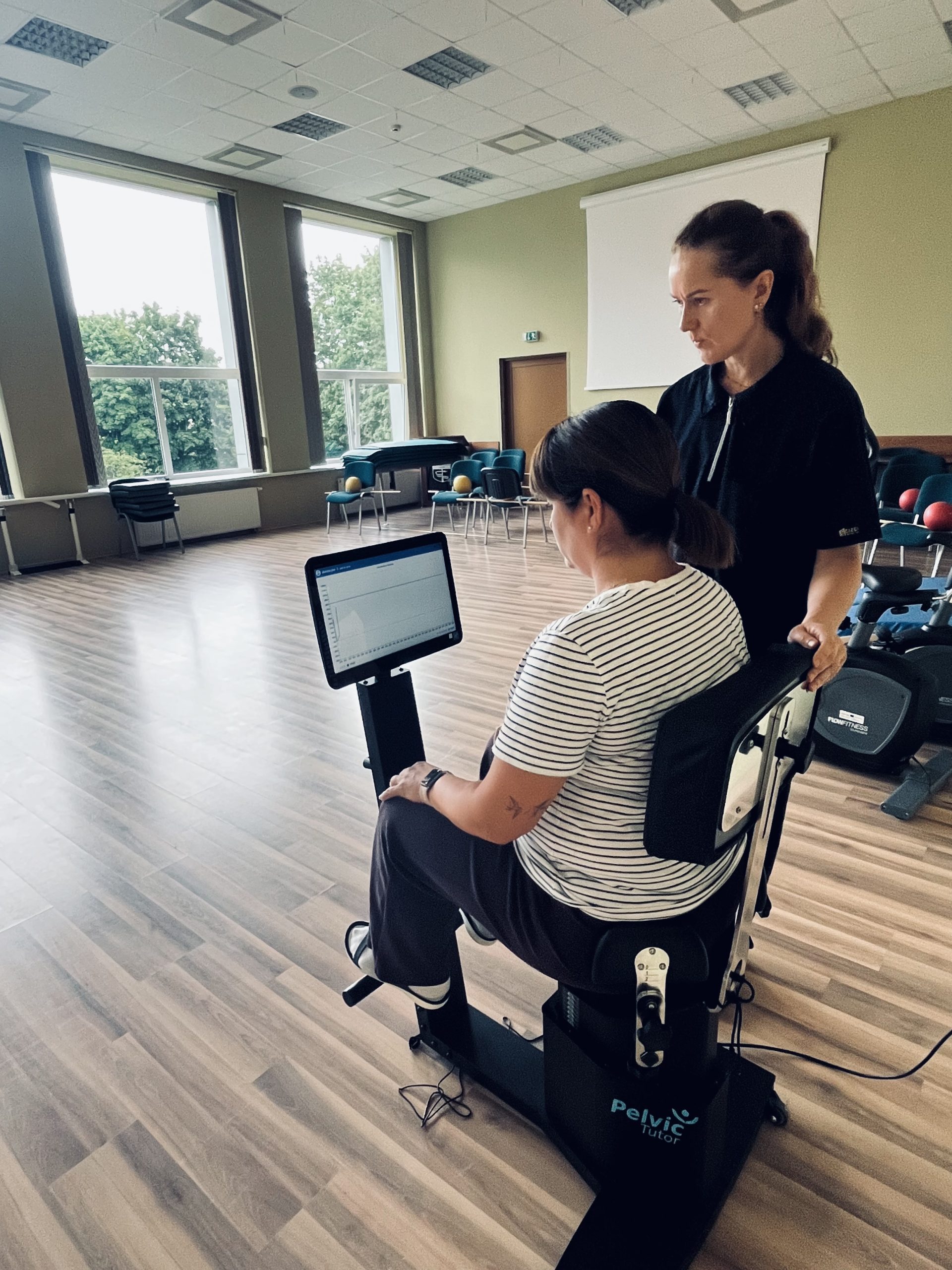 A woman using a Pelvic Tutor device for rehabilitation exercises, guided by a therapist in a spacious, well-lit therapy room. The setup showcases innovative digital technology aimed at enhancing rehabilitation services through the SPEED UP Project.