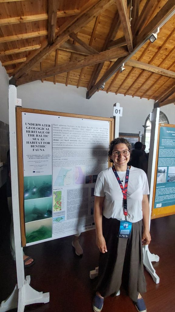 A researcher stands in front of a poster titled "Underwater Geological Heritage of the Baltic Sea," showcasing findings on marine habitats and benthic fauna. The setting features wooden beams and other research presentations in the background.