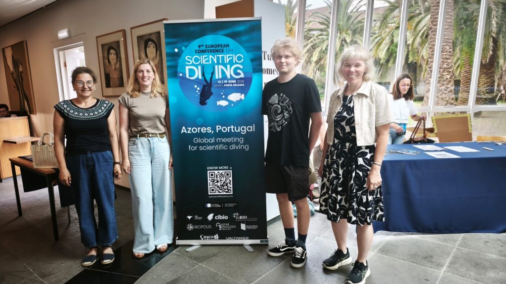 Group of attendees at the 9th European Conference on Scientific Diving in Ponta Delgada, Azores, Portugal, standing next to a promotional banner for the event, showcasing the dates and location.