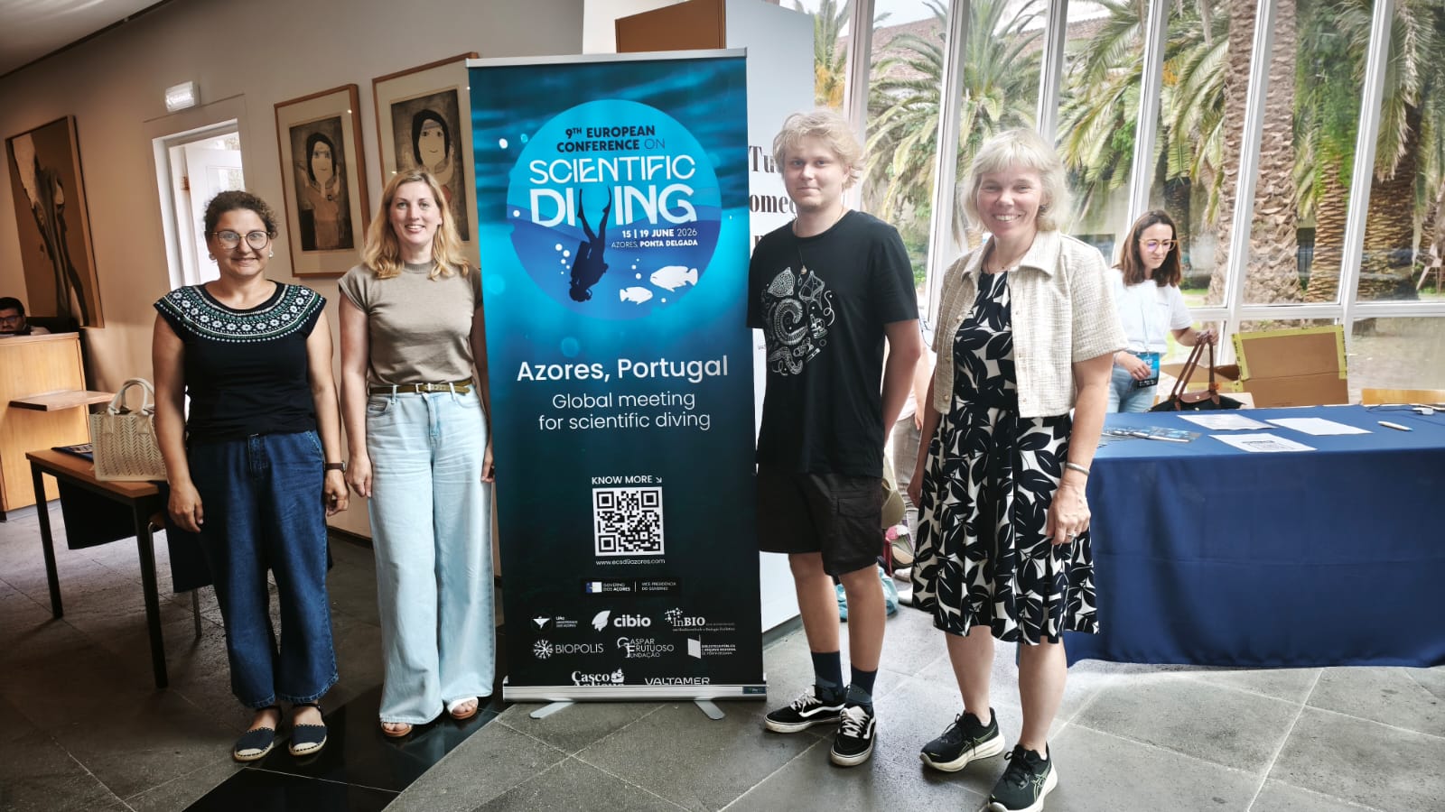 Group of attendees at the 9th European Conference on Scientific Diving in Ponta Delgada, Azores, Portugal, standing next to a promotional banner for the event, showcasing the dates and location.