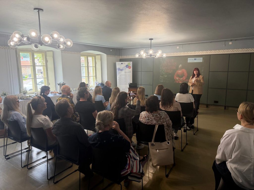 Lecture for tourism professionals at Krustpils Castle, featuring a speaker presenting to an engaged audience seated in a well-lit room. The setting includes windows, a modern chandelier, and informational displays, highlighting the event's focus on tourism and guiding.