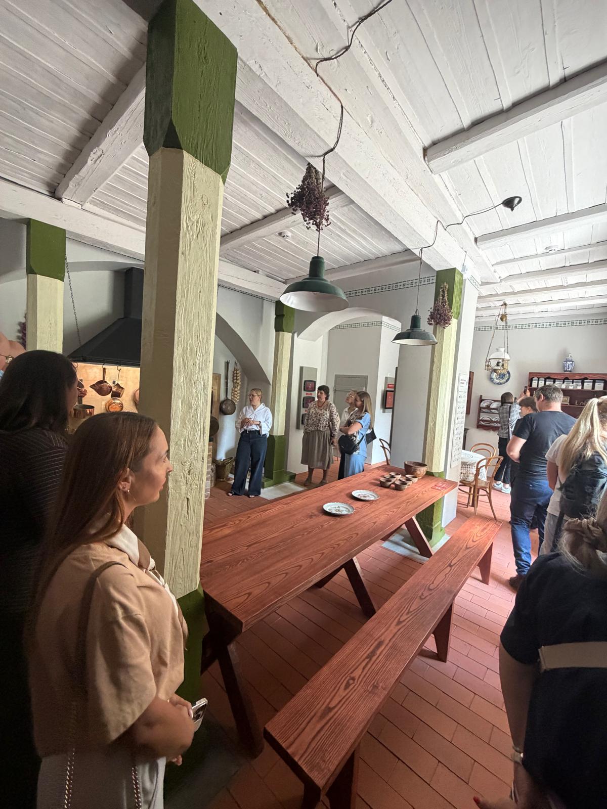 Interior view of a historic kitchen at Krustpils Castle, featuring a large wooden table with plates and traditional decor. Attendees of a tourism lecture are gathered, observing the space, which showcases heritage elements and rustic design.