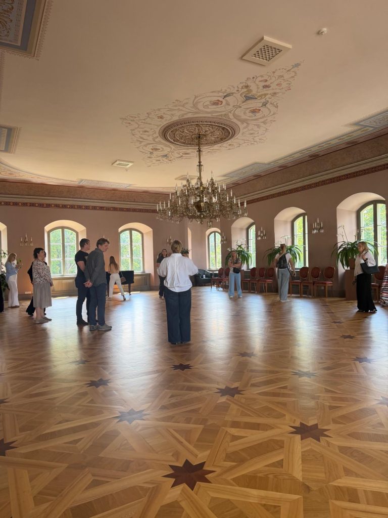 Interior view of a spacious room in Krustpils Castle, featuring a chandelier, decorative ceiling, and wooden floor with intricate patterns. Several individuals engage in a lecture for tourism professionals and guides, surrounded by large windows and elegant furnishings.