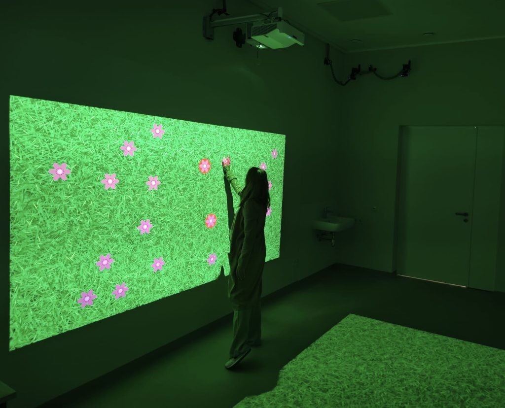 A person interacts with a vibrant digital display featuring a green grass background and colorful flowers, showcasing innovative rehabilitation technology in a modern therapy room as part of the SPEED UP project.