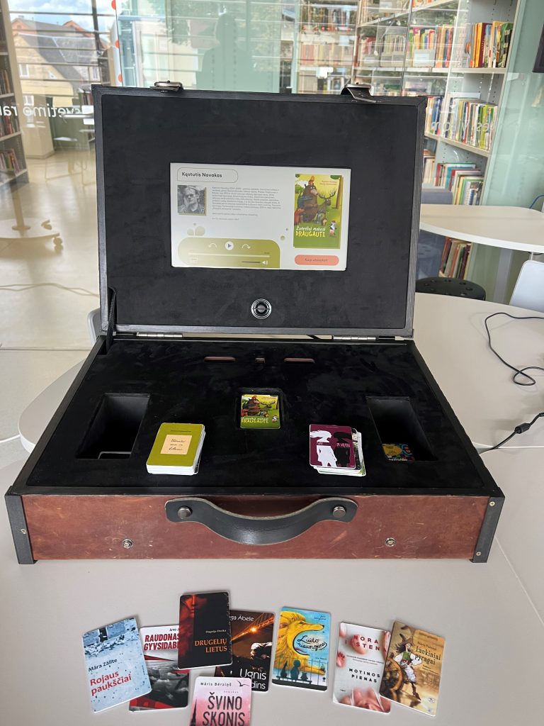 Interactive book suitcase featuring a display with information about author Kęstutis Navakas, surrounded by various small books and cards, designed to enhance the literary experience in a modern library setting. Interactive book suitcase featuring a display with information about author Kęstutis Navakas, surrounded by various small books and cards, designed to enhance the literary experience in a modern library setting.