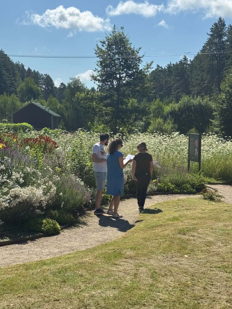 Three individuals engage in a discussion while exploring a vibrant garden filled with diverse plants and flowers, under a clear blue sky. The scene highlights the connection between nature and educational experiences related to the Green Stories Project. Three individuals engage in a discussion while exploring a vibrant garden filled with diverse plants and flowers, under a clear blue sky. The scene highlights the connection between nature and educational experiences related to the Green Stories Project.