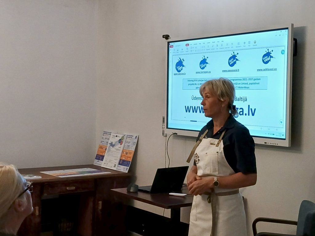 A speaker presents at the Water Tourism Trends seminar in Latgale, held at Lielborne Manor, with informational materials and a digital screen displaying relevant project details in the background.