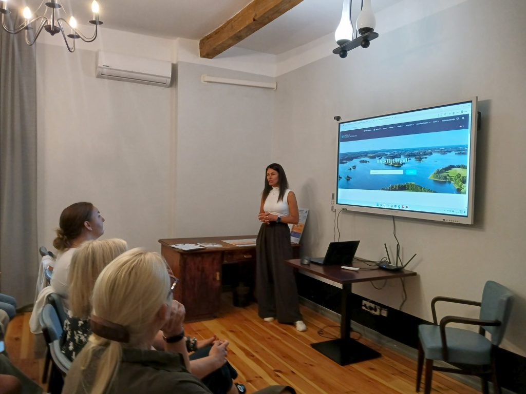Speaker presenting at the "Water Tourism Trends in Latgale" seminar at Lielborne Manor, showcasing a digital presentation on sustainable tourism initiatives, with an audience engaged in discussion.