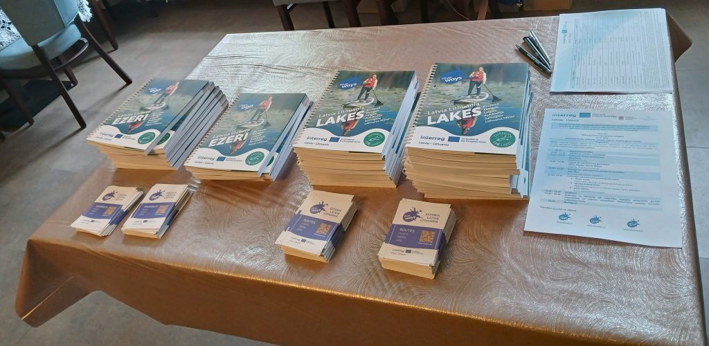 Table displaying promotional materials for the "Water Tourism Trends in Latgale" seminar, featuring stacks of guidebooks about lakes in Latvia and Lithuania, along with brochures and an agenda for the event at Lielborne Manor.