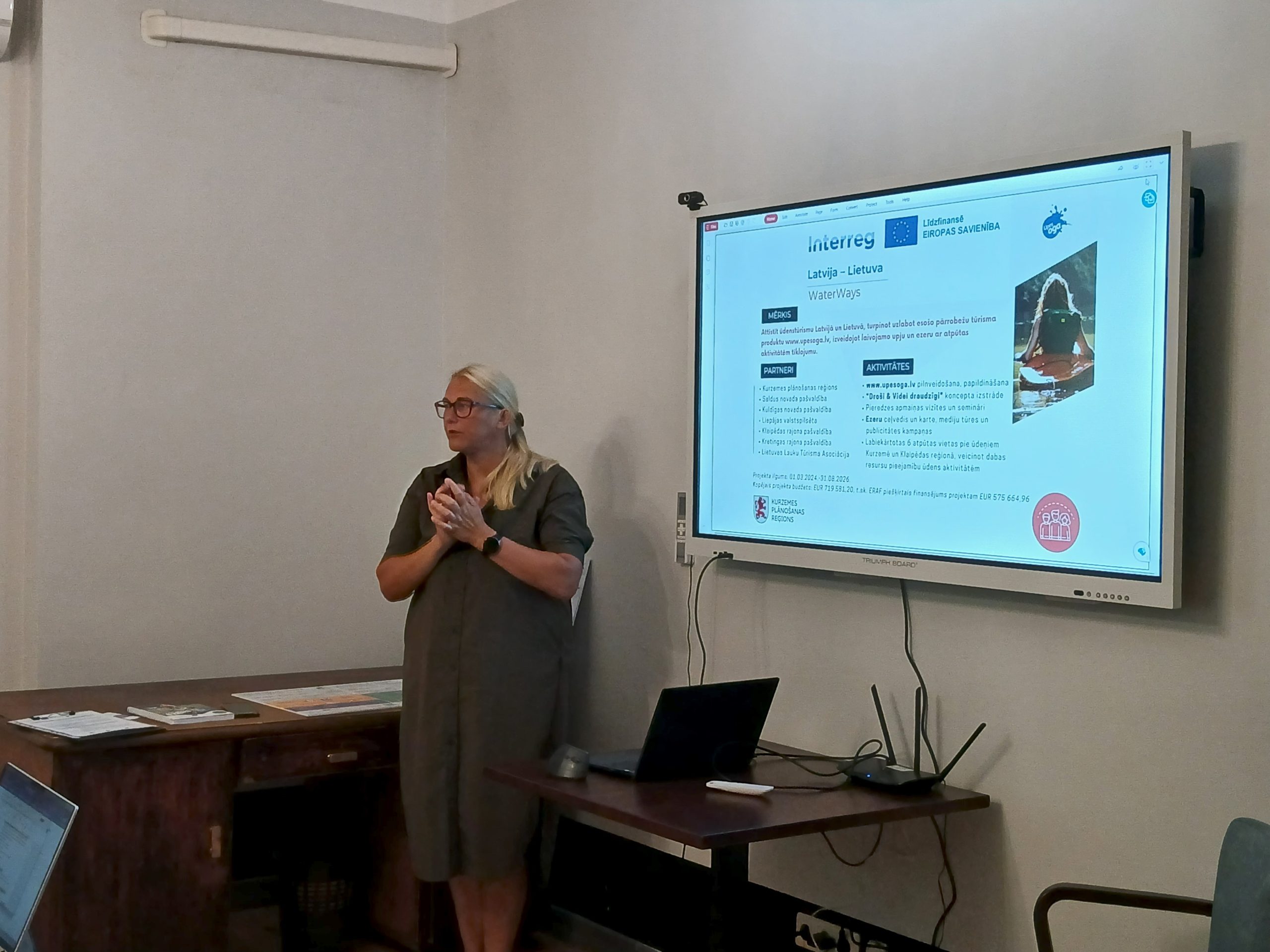 Speaker presenting at the "Water Tourism Trends in Latgale" seminar at Lielborne Manor, with a digital screen displaying information on the WaterWays project. A laptop and printed materials are visible on the table.