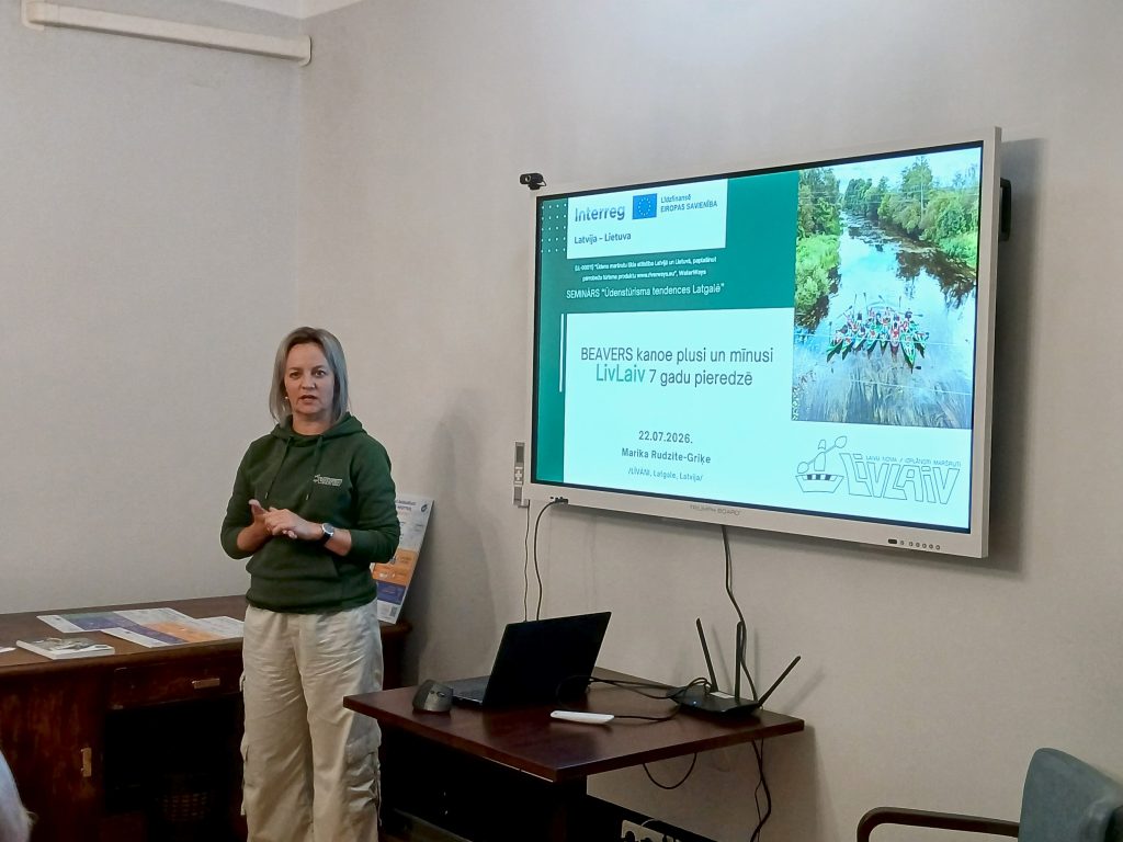 Presenter discussing water tourism trends at a seminar in Lielborne Manor, with a digital screen displaying information about canoeing experiences and a scenic river backdrop.