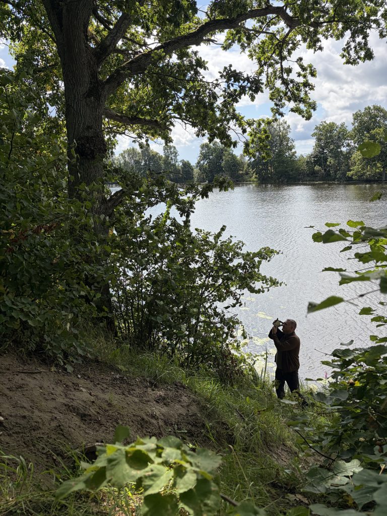 Person photographing geological features near a riverbank in Mažeikiai District, surrounded by lush greenery and trees. The image captures the natural landscape and highlights the area's geological inventory work. Person photographing geological features near a riverbank in Mažeikiai District, surrounded by lush greenery and trees. The image captures the natural landscape and highlights the area's geological inventory work.