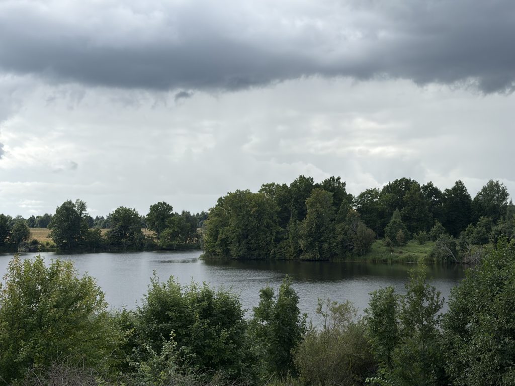 Scenic view of a tranquil lake surrounded by lush greenery and trees under a cloudy sky in Mažeikiai District, highlighting the area's geological features and natural landscape. Scenic view of a tranquil lake surrounded by lush greenery and trees under a cloudy sky in Mažeikiai District, highlighting the area's geological features and natural landscape.