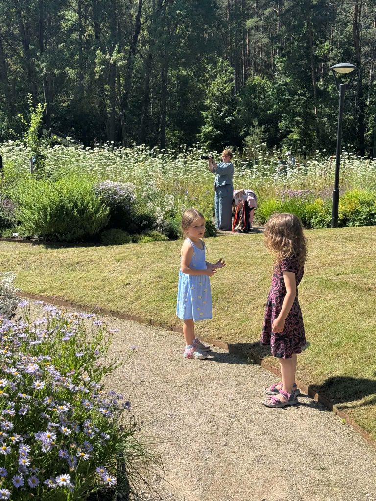 Two young girls interact in a lush garden filled with wildflowers, while an adult in the background captures the moment with a camera. The image highlights the theme of nature exploration in educational settings, reflecting the Green Stories Project's focus on immersive learning experiences. Two young girls interact in a lush garden filled with wildflowers, while an adult in the background captures the moment with a camera. The image highlights the theme of nature exploration in educational settings, reflecting the Green Stories Project's focus on immersive learning experiences.